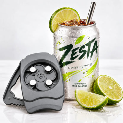 A gray silicone carbonation release tool with metal release pins is shown next to a ZESTA sparkling water can topped with a lime wheel and straw, with lime slices on a white surface.